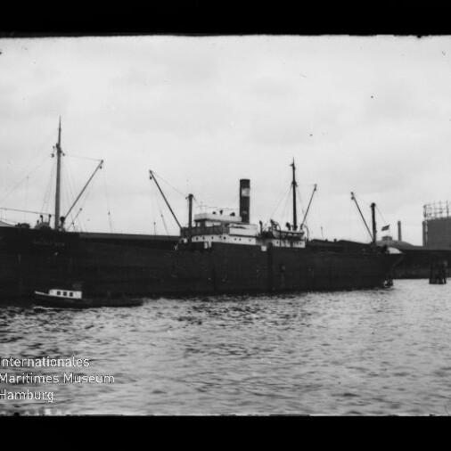 Schwarzweißaufnahme eines großen Frachtschiffs mit Schornstein und Ladebaum-Masten, an einem Kai liegend, kleines Boot am Bug, Gasholder im Hintergrund und leicht bewegte Wasseroberfläche im Vordergrund.