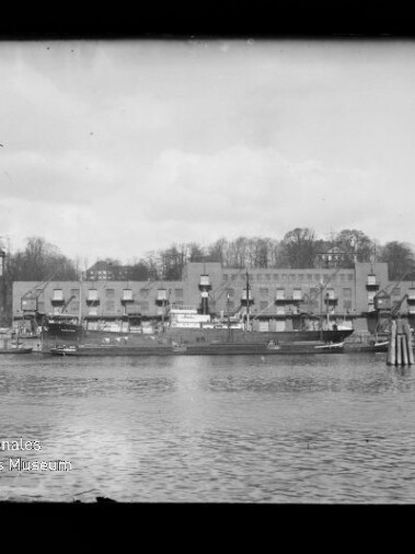 Blick über das Wasser auf einen Hafen mit mehreren angelegten Frachtschiffen vor einer langen Reihe alter Lagerhäuser und Gebäuden, im Hintergrund ein baumbewachsener Hang.