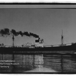 Schwarz-weiße Seitenaufnahme eines Frachtschiffs/Dampfers auf ruhigem Wasser mit hohem Schornstein, aus dem dunkler Rauch aufsteigt, und deutlicher Spiegelung im Wasser; unten links Stempel des Internationalen Maritimen Museums Hamburg.