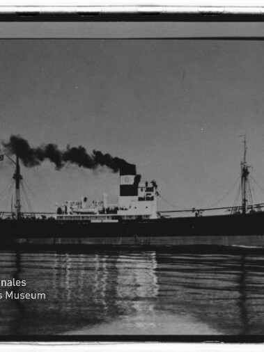 Schwarz-weiße Seitenaufnahme eines Frachtschiffs/Dampfers auf ruhigem Wasser mit hohem Schornstein, aus dem dunkler Rauch aufsteigt, und deutlicher Spiegelung im Wasser; unten links Stempel des Internationalen Maritimen Museums Hamburg.