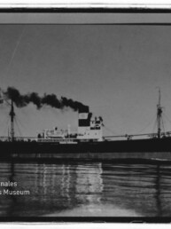 Seitliche Aufnahme eines Dampffrachters, der dichten schwarzen Rauch aus dem Schornstein ausstößt; das Schiff fährt über ruhiges Wasser mit klarer Spiegelung, links unten ist das Logo des Internationalen Maritimen Museums Hamburg sichtbar.