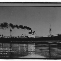 Seitliche Aufnahme eines Dampffrachters, der dichten schwarzen Rauch aus dem Schornstein ausstößt; das Schiff fährt über ruhiges Wasser mit klarer Spiegelung, links unten ist das Logo des Internationalen Maritimen Museums Hamburg sichtbar.
