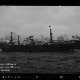 Schwarzweiß-Foto eines Frachtschiffs im Hafen mit rauchendem Schornstein, Kränen und weiteren Schiffen im Hintergrund, bewegter See und Museum-Logo in der unteren linken Ecke.
