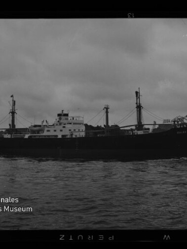 Schwarzweiß-Foto eines Frachtschiffs in Seitenansicht im Hafen bei bewölktem Himmel, mit Aufbau und Ladebäumen, hinter dem Schiff mehrere Dockkräne an der Kaikante und ruhiges Wasser im Vordergrund; links unten das Logo des Internationalen Maritimen Museums Hamburg sichtbar.