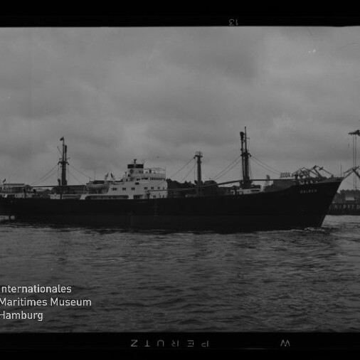 Schwarzweiß-Foto eines Frachtschiffs in Seitenansicht im Hafen bei bewölktem Himmel, mit Aufbau und Ladebäumen, hinter dem Schiff mehrere Dockkräne an der Kaikante und ruhiges Wasser im Vordergrund; links unten das Logo des Internationalen Maritimen Museums Hamburg sichtbar.
