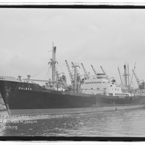 Schwarzweiß-Foto eines Frachtschiffs mit dem gut lesbaren Namen DALBEK am Bug, das am Hafen liegt, mit mehreren Ladebäumen und Hafenkränen im Hintergrund, ruhigem Wasser im Vordergrund und Museumslogo unten links.