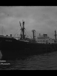Schwarzweiß-Foto eines großen Frachtschiffs mit dem Namen DALBEK, längsseits am Kai liegend mit mehreren Masten und Ladekränen, dunklem Wasser und bewölktem Himmel; links unten sichtbar das Logo des Internationalen Maritimen Museums Hamburg.