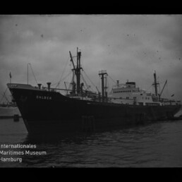Schwarzweiß-Foto eines großen Frachtschiffs mit dem Namen DALBEK, längsseits am Kai liegend mit mehreren Masten und Ladekränen, dunklem Wasser und bewölktem Himmel; links unten sichtbar das Logo des Internationalen Maritimen Museums Hamburg.