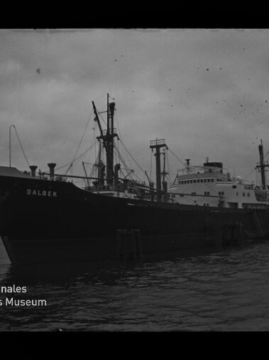 Schwarzweiß-Foto eines großen Frachtschiffs mit dem Namen DALBEK, längsseits am Kai liegend mit mehreren Masten und Ladekränen, dunklem Wasser und bewölktem Himmel; links unten sichtbar das Logo des Internationalen Maritimen Museums Hamburg.