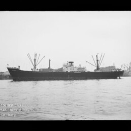Schwarzweißaufnahme eines Frachtschiffs mit mehreren Ladebäumen und markanter Aufbauten, seitlich im Hafen auf ruhiger See, mit Kaianlagen und Hafenkränen im Hintergrund sowie Museumslogo unten links.