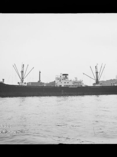 Schwarzweißaufnahme eines Frachtschiffs mit mehreren Ladebäumen und markanter Aufbauten, seitlich im Hafen auf ruhiger See, mit Kaianlagen und Hafenkränen im Hintergrund sowie Museumslogo unten links.