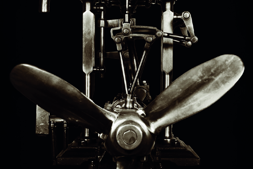 Polierter dreiblättriger Propeller vor freiliegendem, mechanischem Metalltriebwerk auf schwarzem Hintergrund.