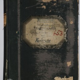 Abgenutztes dunkelgrünes bis schwarzes Buch mit stark abgegriffenem Einband und beschädigten Kanten; mittig ein achteckiges, vergilbtes Etikett mit verblasstem Druck, handschriftlicher Ortsangabe 