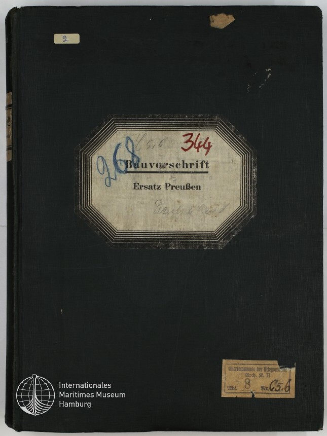 Schwarzer Leinen-Einband eines Hefts mit abgenutzten Ecken, mittig ein achteckiges Etikett mit dem gedruckten Titel Bauvorschrift Ersatz Preußen und handschriftlichen Zahlen in Blau und Rot, sowie kleinen Papieretiketten oben links und unten rechts und dem Logo des Internationalen Maritimen Museums Hamburg unten links.