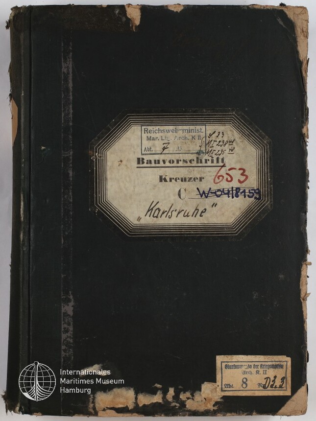 Vorderseite eines stark abgenutzten schwarzen Buchbandes mit einem zentralen achteckigen Etikett, auf dem „Bauvorschrift Kreuzer ‚Karlsruhe‘“ sowie handschriftliche Notizen und rote und blaue Stempelnummern zu sehen sind, dazu ein kleines Etikett unten rechts und das Logo des Internationalen Maritimen Museums Hamburg unten links.
