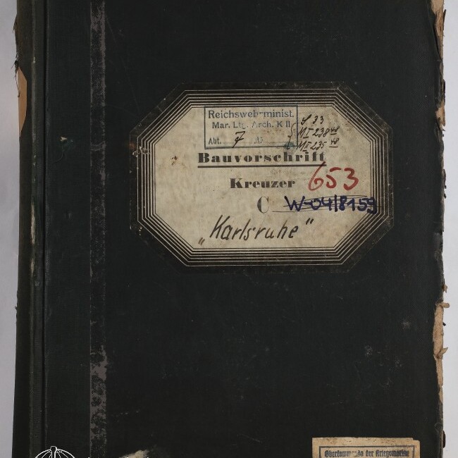 Vorderseite eines stark abgenutzten schwarzen Buchbandes mit einem zentralen achteckigen Etikett, auf dem „Bauvorschrift Kreuzer ‚Karlsruhe‘“ sowie handschriftliche Notizen und rote und blaue Stempelnummern zu sehen sind, dazu ein kleines Etikett unten rechts und das Logo des Internationalen Maritimen Museums Hamburg unten links.
