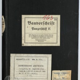 Schwarzer Leineneinband eines Ordners mit mittig aufgeklebtem Schild 'Bauvorschrift Panzerschiff E' (oben handschriftlich rot die Ziffer 163), darunter links ein größeres russischsprachiges Etikett und rechts unten ein kleines Papieretikett.