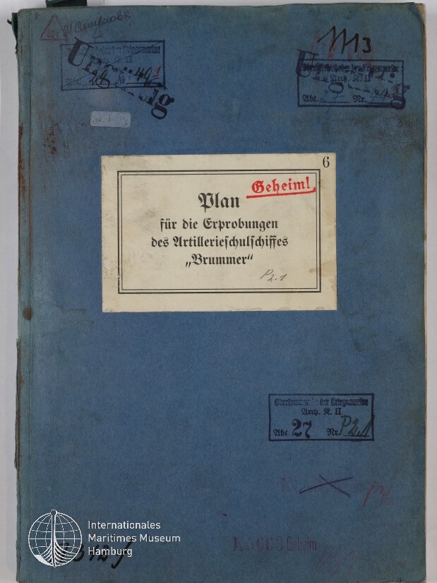 Blaues, altes Ordnerdeckblatt mit einem aufgeklebt weißen Etikett, auf dem in schwarzer Schrift und rot gestempelt „Geheim! Plan für die Erprobungen des Artillerieschulschiffes ‚Brummer‘“ steht; Umschlag zeigt mehrere Stempel, handschriftliche Notizen, Nummern, Gebrauchsspuren und ein Museumssignet unten links.