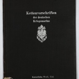 Schwarzer Leineneinband eines Buches mit silberner Frakturschrift ‚Kettenvorschriften der deutschen Kriegsmarine‘, einem Reichsadler-Emblem in der Mitte, dem Vermerk ‚Kaiserliche Werft, Kiel‘ unten, leichter Eckenabnutzung und dem Logo des Internationalen Maritimen Museums Hamburg in der unteren linken Ecke.