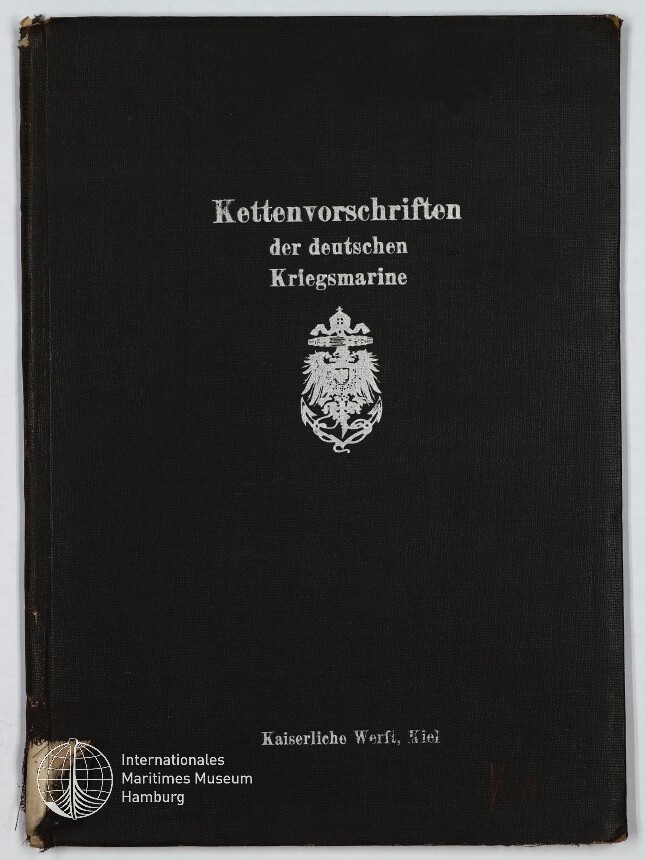 Schwarzer Leineneinband eines Buches mit silberner Frakturschrift ‚Kettenvorschriften der deutschen Kriegsmarine‘, einem Reichsadler-Emblem in der Mitte, dem Vermerk ‚Kaiserliche Werft, Kiel‘ unten, leichter Eckenabnutzung und dem Logo des Internationalen Maritimen Museums Hamburg in der unteren linken Ecke.
