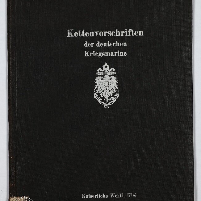 Schwarzer Leineneinband eines Buches mit silberner Frakturschrift ‚Kettenvorschriften der deutschen Kriegsmarine‘, einem Reichsadler-Emblem in der Mitte, dem Vermerk ‚Kaiserliche Werft, Kiel‘ unten, leichter Eckenabnutzung und dem Logo des Internationalen Maritimen Museums Hamburg in der unteren linken Ecke.