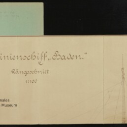 Auf einem aufgeklappten beigefarbenen Planblatt mit sichtbaren Falz- und Lochungen links steht in kalligrafischer Schrift „S.M. Linienschiff ‚Baden.‘ Längsschnitt 1:100“, rechts ist eine feine Bleistiftskizze eines Mastes mit Wanten zu sehen; links liegt ein blassgrünes Registerblatt und unten links prangt das Logo des Internationalen Maritimen Museums Hamburg.