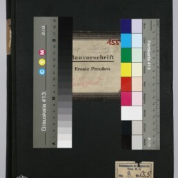 Schwarz bezogenes Buch mit mittig aufgeklebstem Etikett „Bauvorschrift Ersatz Preußen“, zwei Farbkarten/Grauskalen links und rechts, kleinem Sammlungsetikett unten rechts, Logodesign des Internationalen Maritimen Museums Hamburg unten links und einem Lineal/Maßstab am unteren Bildrand.