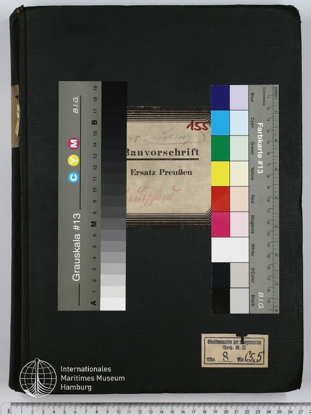 Schwarz bezogenes Buch mit mittig aufgeklebstem Etikett „Bauvorschrift Ersatz Preußen“, zwei Farbkarten/Grauskalen links und rechts, kleinem Sammlungsetikett unten rechts, Logodesign des Internationalen Maritimen Museums Hamburg unten links und einem Lineal/Maßstab am unteren Bildrand.