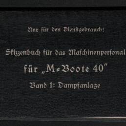 Schwarzes, quer liegendes Skizzenbuch mit weißer Frakturschrift: „Nur für den Dienstgebrauch! Skizzenbuch für das Maschinenpersonal für ‚M‑Boote 40‘ Band 1: Dampfanlage“, oben rechts der Schriftzug „Ausfertigung 876“, handschriftliches Etikett oben links sowie links unten das Logo des Internationalen Maritimen Museums Hamburg und rechts unten das Logo der Deutschen Werke Kiel AG.