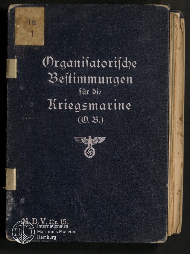 Dunkelblauer Einband eines Buchs mit weißer Frakturaufschrift „Organisatorische Bestimmungen für die Kriegsmarine (O.B.)“, darunter ein Reichsadler mit Hakenkreuz und unten links das Logo des Internationalen Maritimen Museums Hamburg.