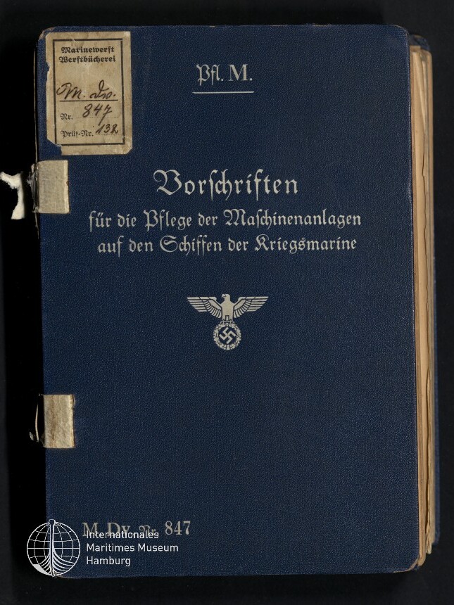 Dunkelblaues Einbandbuch mit silberner Frakturschrift 'Vorschriften für die Pflege der Maschinenanlagen auf den Schiffen der Kriegsmarine', oben die Abkürzung 'Pfl. M.', in der Mitte ein Reichsadler mit Hakenkreuz sowie links oben kleine Etiketten und unten links das Signet des Internationalen Maritimen Museums Hamburg.
