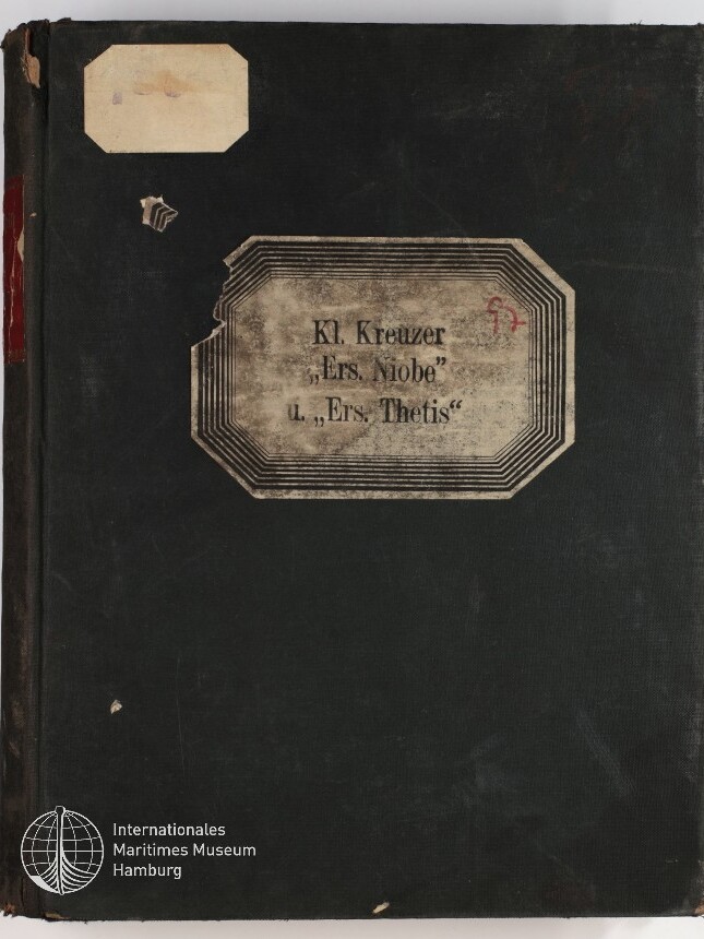 Schwarzer, abgenutzter Buch- oder Ordnerdeckel mit vergilbtem, achteckigem Etikett in der Mitte mit der Aufschrift Kl. Kreuzer „Ers. Niobe“ L. „Ers. Thetis“, kleinem abgelösten Etikett oben links und weißem Logo des Internationalen Maritimen Museums Hamburg unten links