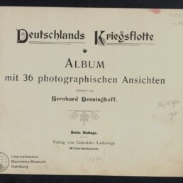 Titelblatt eines Albums mit der Überschrift „Deutschlands Kriegsflotte“ in dekorativer Frakturschrift, darunter „Album mit 36 photographischen Ansichten“, Erläuterung von Bernhard Denninghoff, Verlagsangabe, dekoratives Ornament, Stempel des Internationalen Maritimen Museums Hamburg und mehrere handschriftliche Notizen am Rand.