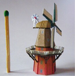 kleines Modell einer holländischen Windmühle aus Pappe mit umlaufender Galerie und vier Flügeln, daneben ein Streichholz zum Größenvergleich