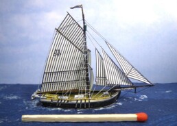 Kleines Modell eines Segelschiffs mit gestreiften Segeln und sichtbaren Takelagen, im Vordergrund ein Streichholz zur Größendarstellung.