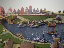 Modellhafen mit zahlreichen kleinen Fischerbooten an Kaimauer, grüner Uferbereich und Reihenhausfassaden im Hintergrund.