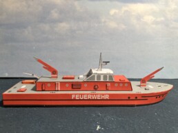 Rotes Modellboot mit Aufschrift FEUERWEHR, Aufbauten, Rettungsring und ausklappbaren roten Auslegern auf grauem Deck.