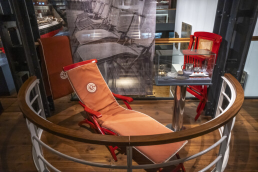 Roter Liegestuhl mit orangefarbenem Polster in einer Museumspr&auml;sentation, daneben eine Glasvitrine mit kleinem Tee-Service.