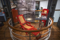 Zwei rote Klapp-Liegestühle mit geflochtenen Sitzflächen neben einem kleinen Vitrinentisch auf einer Museumsbalkon-Galerie.