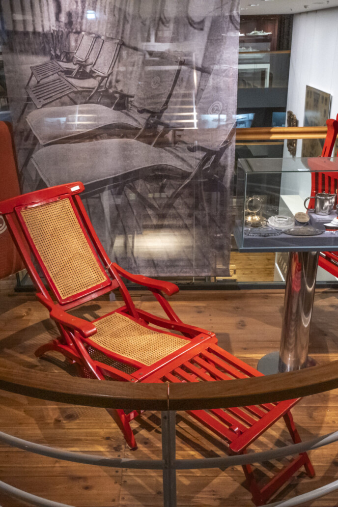 Zwei rote Klapp-Liegestühle mit geflochtenen Sitzflächen neben einem kleinen Vitrinentisch auf einer Museumsbalkon-Galerie.