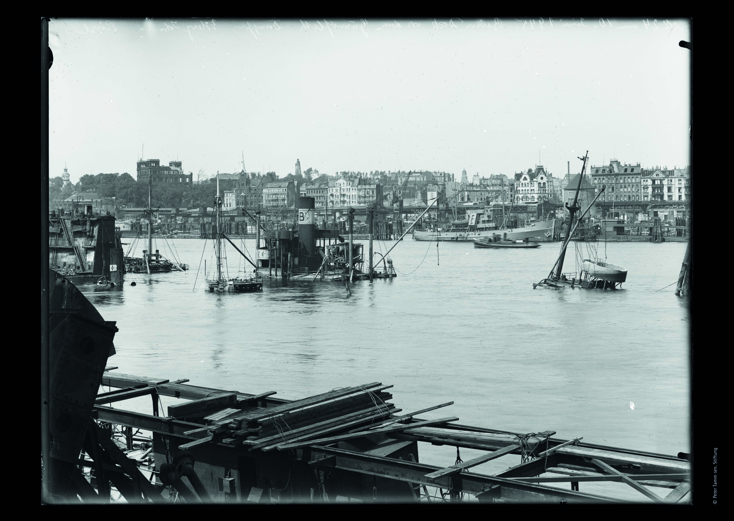 Hafenblick: Bauholz und Ger&uuml;st im Vordergrund, halb versunkene Boote und Arbeitsplattformen im Wasser, Stadtsilhouette im Hintergrund.