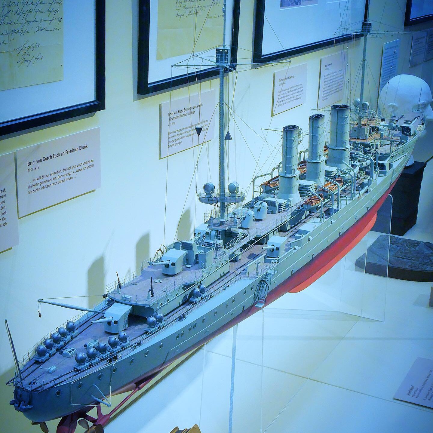 Detailliertes Modell eines Kriegsschiffs mit drei Schornsteinen, Masten und Takelage in Museumsvitrine vor gerahmten Dokumenten.