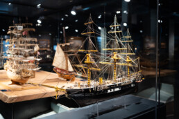 Detailliertes Segelschiffsmodell mit schwarzem Rumpf und gelben Masten in einer Vitrine, weitere Modelle im Hintergrund.