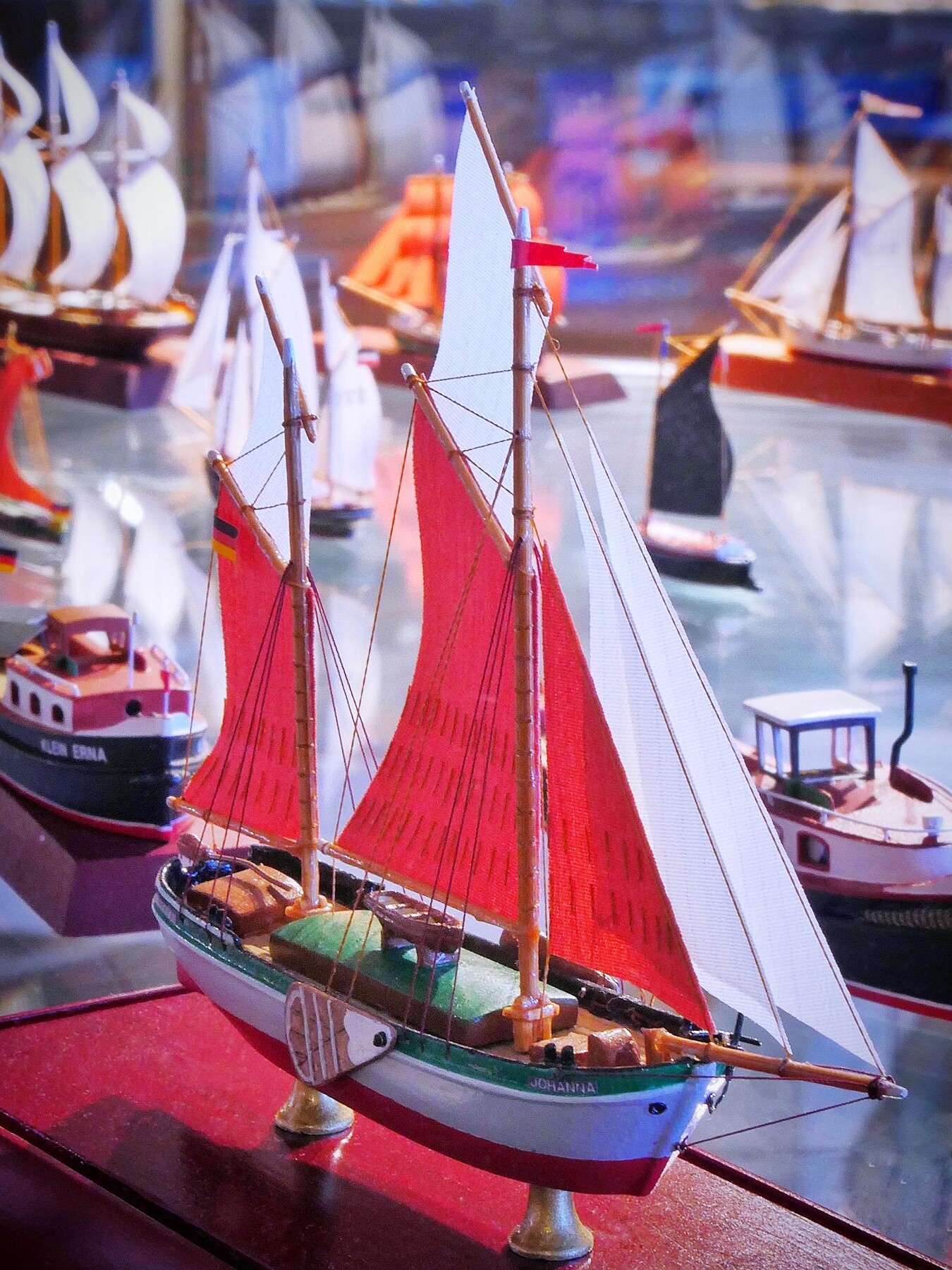 Modellsegelschiff mit roten und weißen Segeln auf Ausstellungsfläche, im Hintergrund mehrere weitere Miniaturboote.