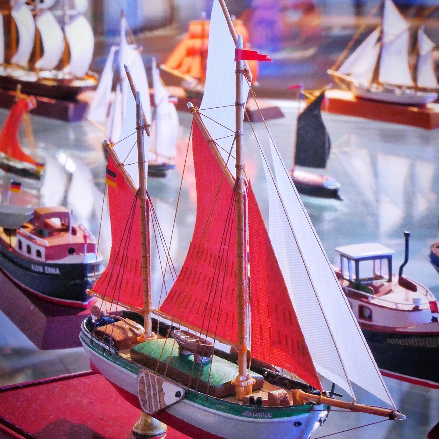 Modellsegelschiff mit roten und weißen Segeln auf Ausstellungsfläche, im Hintergrund mehrere weitere Miniaturboote.