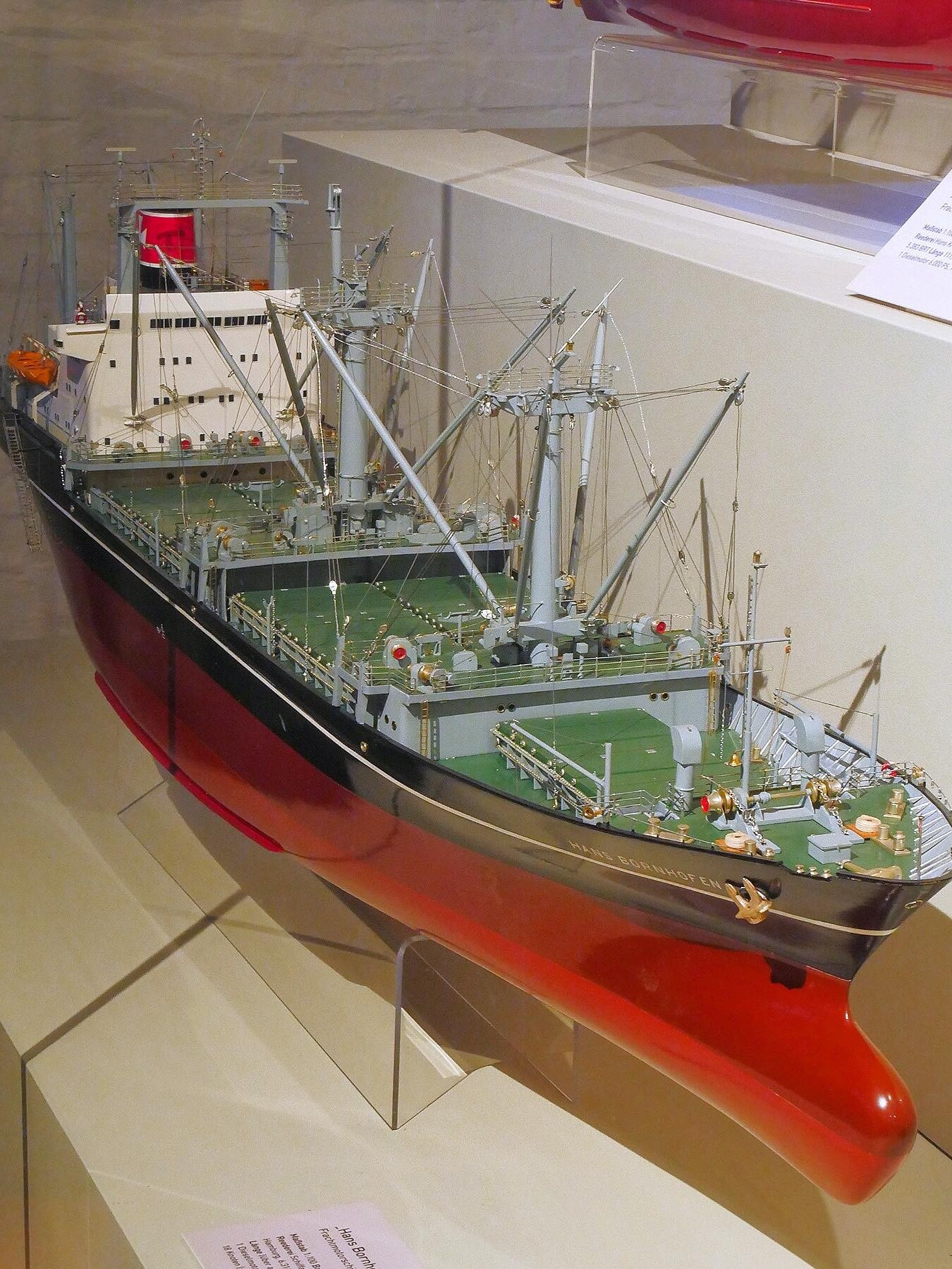 detailliertes Schiffsmodell mit grünem Deck, mehreren Ladekränen und rotem Rumpf in Museumsvitrine.