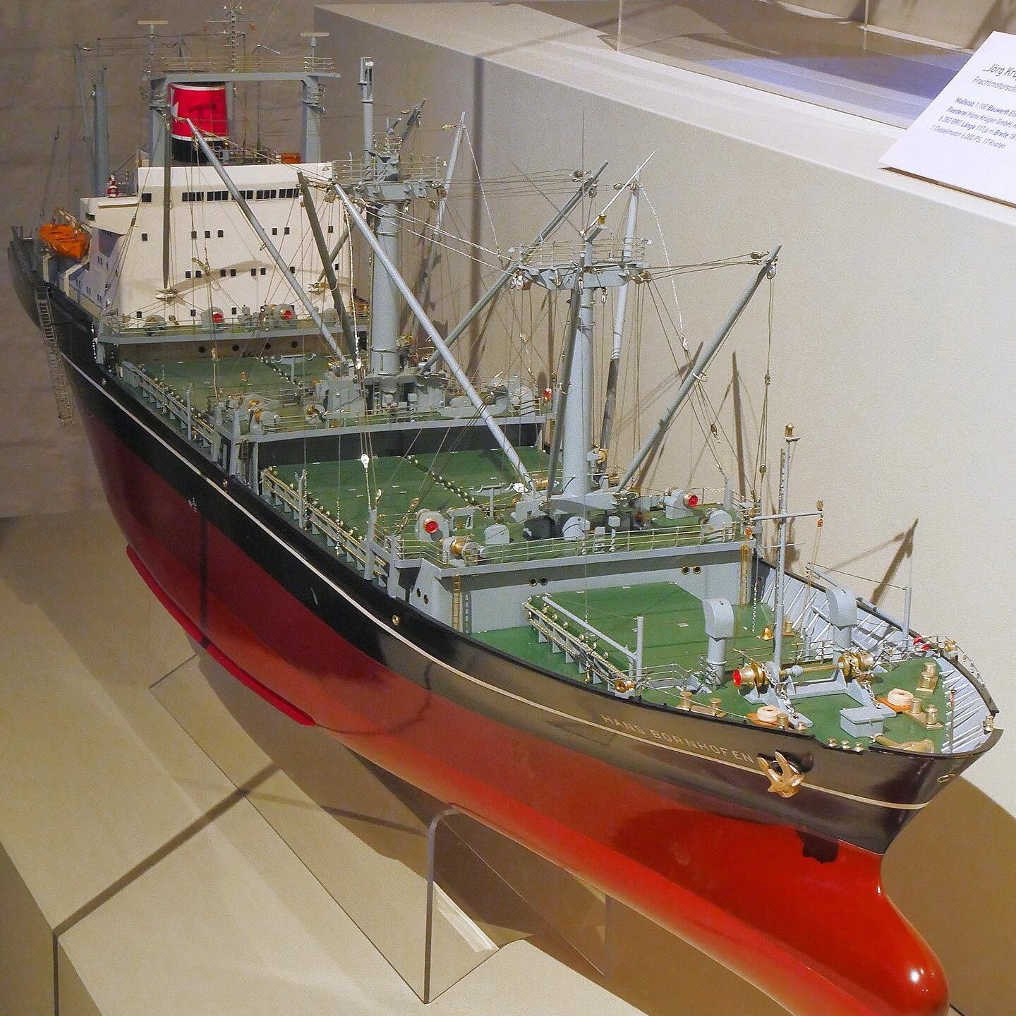 detailliertes Schiffsmodell mit grünem Deck, mehreren Ladekränen und rotem Rumpf in Museumsvitrine.