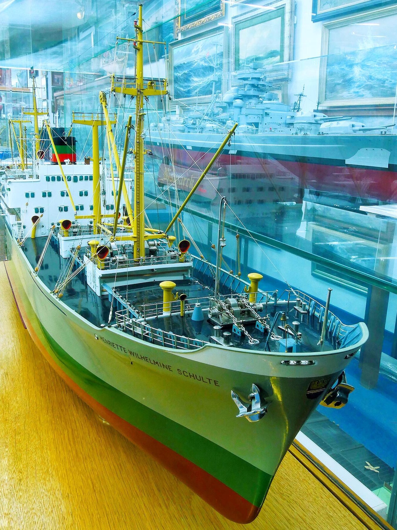 Modell eines grünen Frachtschiffs mit gelben Masten und vielen Takelagen in einer Glasvitrine, weitere Schiffsmodelle und maritime Gemälde im Hintergrund