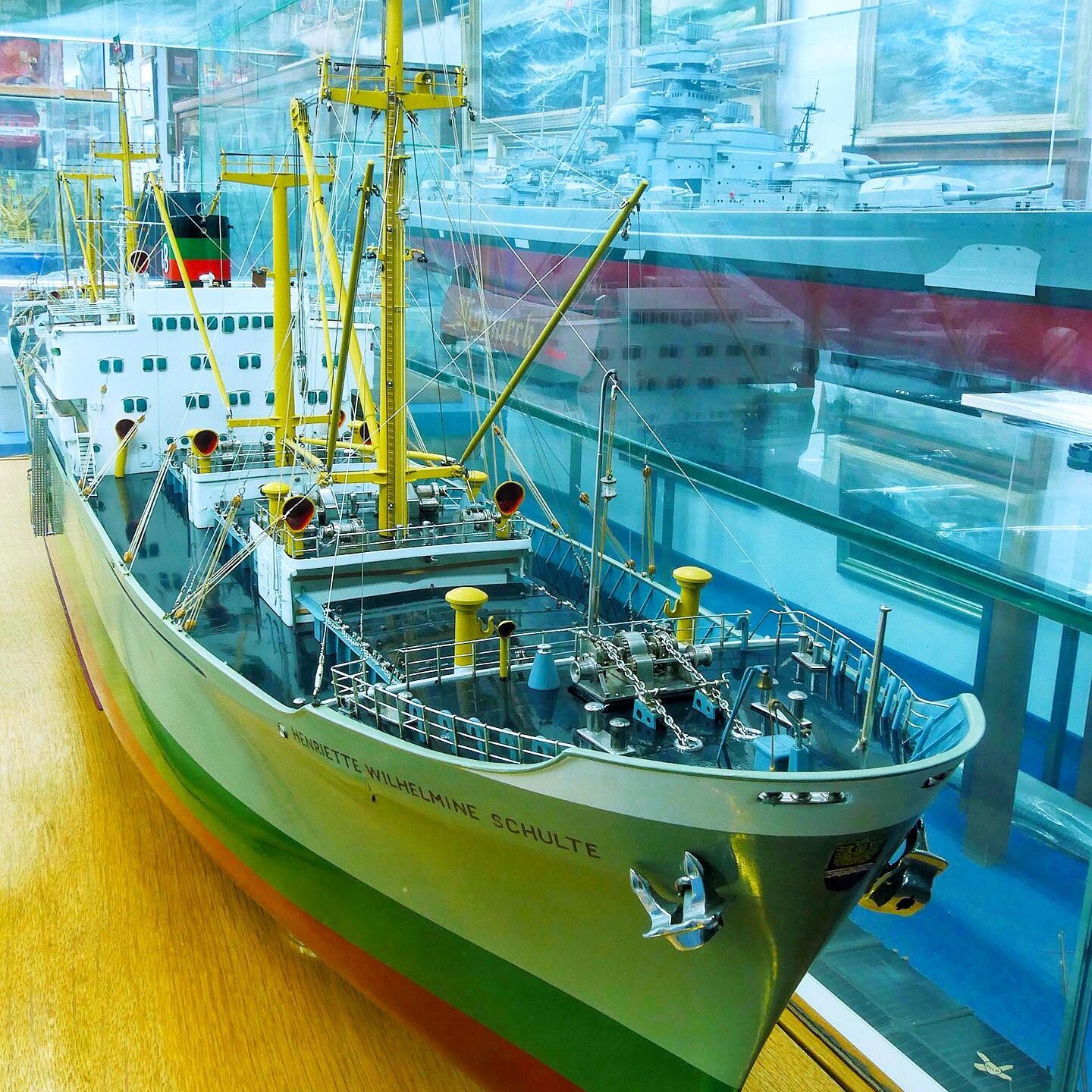 Modell eines grünen Frachtschiffs mit gelben Masten und vielen Takelagen in einer Glasvitrine, weitere Schiffsmodelle und maritime Gemälde im Hintergrund