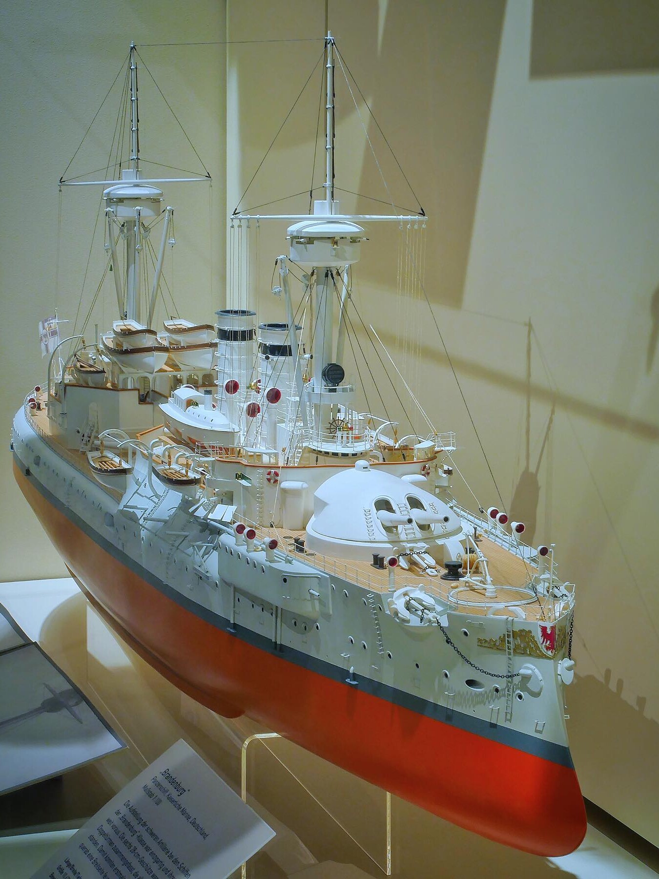 Detailliertes Modell eines Kriegsschiffs mit rotem Rumpf, weißen Aufbauten, Masten, Takelage und Rettungsbooten in einer Vitrine.